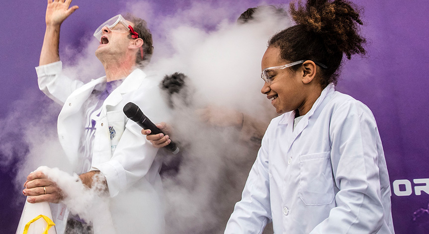 Mad scientist holding a microphone behind a cloud of smoke in between a girl and mad scientist both in lab coats