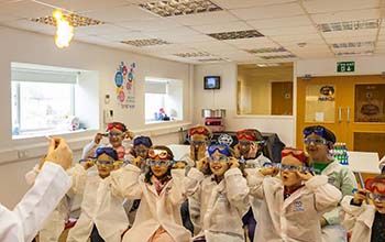 A Mad Scientist throwing lit flash paper in the air in front of kids in class wearing lab coats and safety goggles