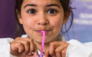 Girl in lab coat blowing up purple balloon using a straw